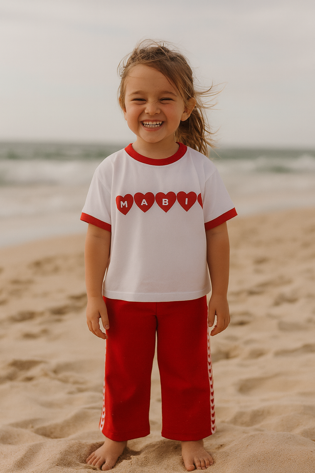 Child wearing a white shirt with red hearts and red pants on a beach.