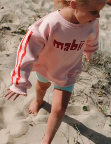 Child wearing a pink sweatshirt with red stripes and 'mabii' text on a sandy background