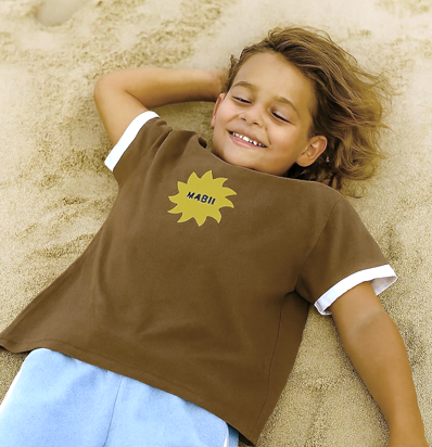 Child wearing a brown shirt with a yellow sun design lying on sand