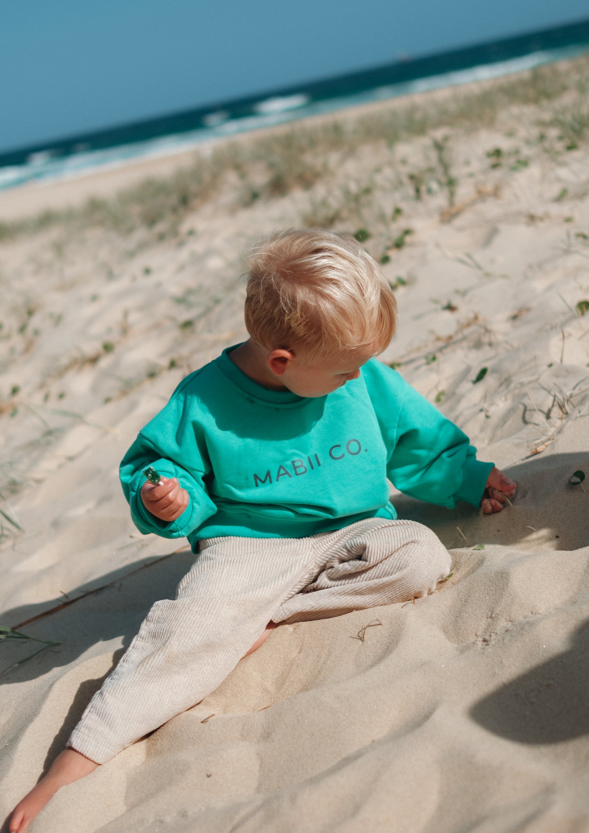 Child sitting on a sandy beach wearing a teal sweatshirt with 'MABII' printed on it.