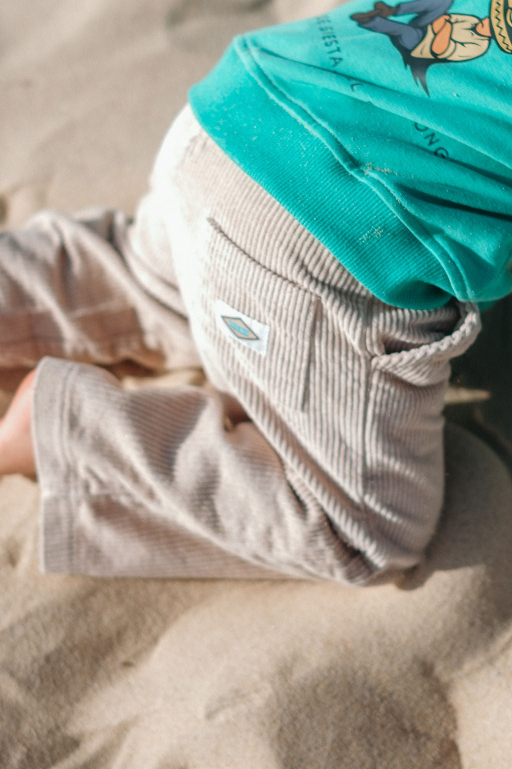 Close-up of a child wearing soft cord beige pants and a green shirt on sand.